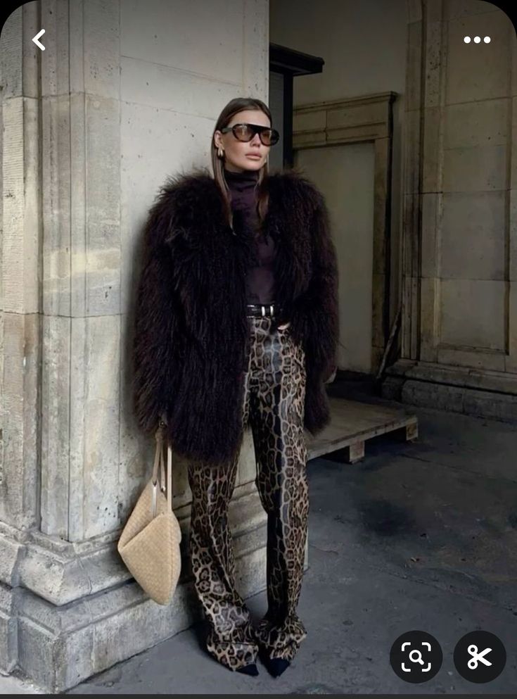 a woman leaning against a pillar wearing leopard print pants and a fur coat with her hands in her pockets