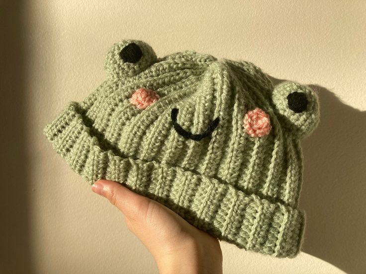 a hand holding up a green knitted frog hat with eyes and nose on it