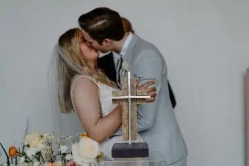 a bride and groom kissing in front of a wooden cross on a table with flowers