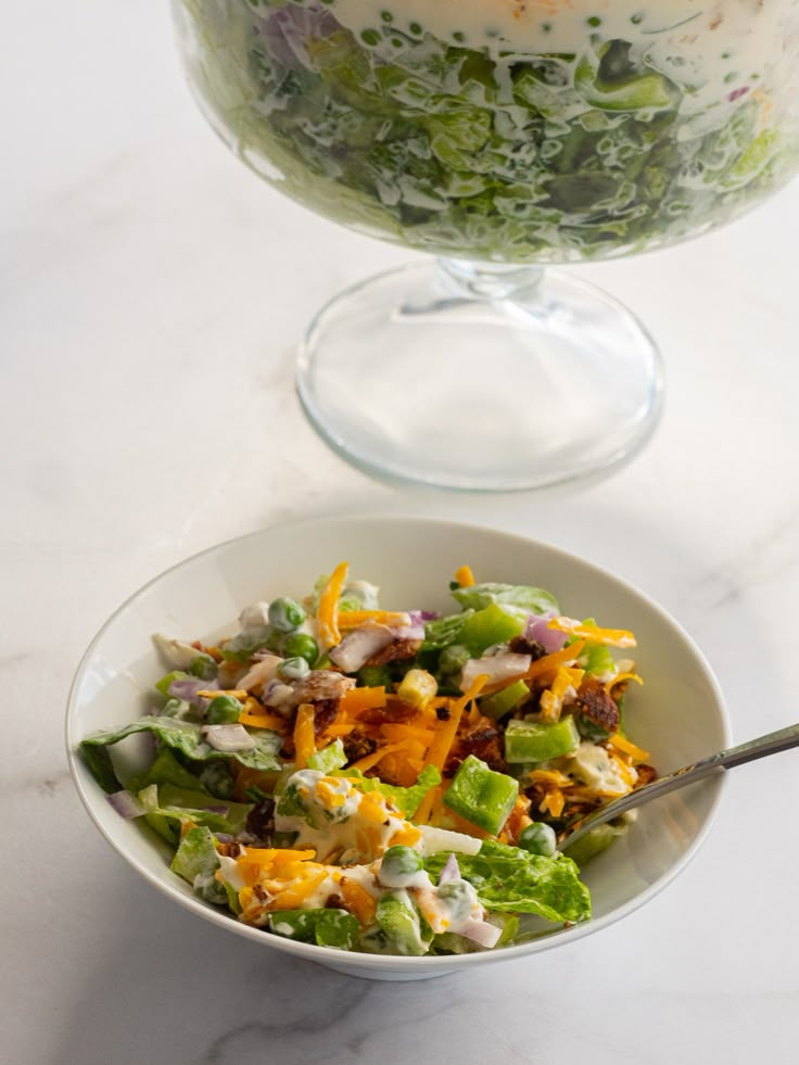 a salad in a bowl next to a large glass cake dish on a marble table