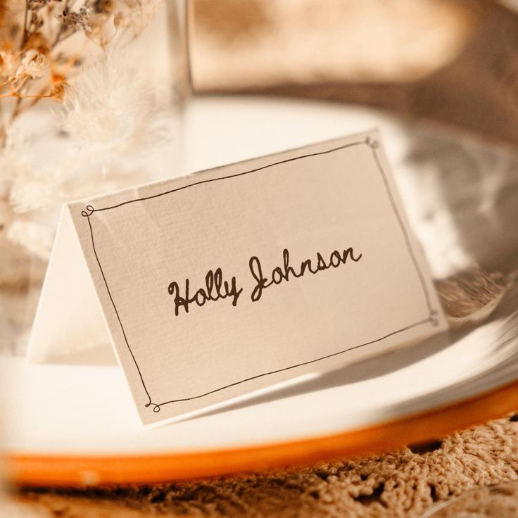 a place card sitting on top of a white plate next to a glass filled with flowers