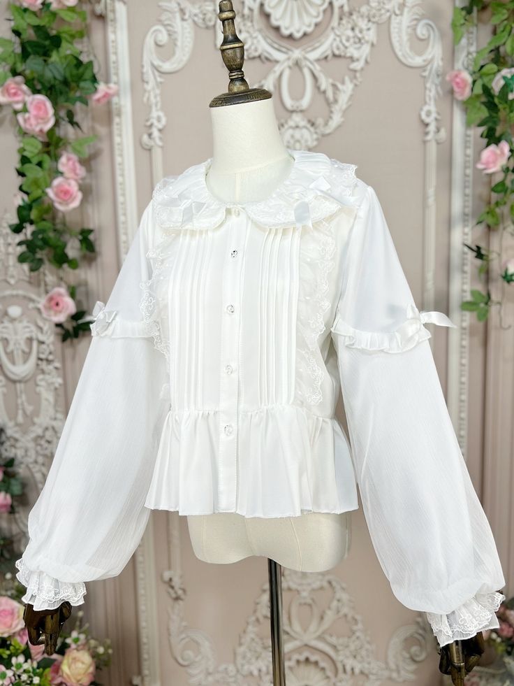 Dress up in this incredibly sweet top that exudes a doll-like charm. The floral bud collar is adorned with delicate lace and a cute bow, adding a touch of elegance. The sleeves feature a unique double-layer design, with a soft fabric layer on the inside and a crinkled chiffon layer on the outside, creating a lovely and whimsical look. The elastic cuffs ensure a comfortable fit, while the irremovable bows on both sides of the sleeves add a playful and adorable touch.  Please note that the price i White Lace Trim Coquettish Top, White Lace Trim Top Feminine Style, Cute Spring Blouse With Bow, White Feminine Lace Top With Puff Sleeves, Cute White Blouse With Doll Collar, Cute White Top With Lace Collar, Cute White Tops With Lace Collar, White Blouse With Doll Collar In Cute Style, White Peter Pan Collar Top With Lace Cuffs