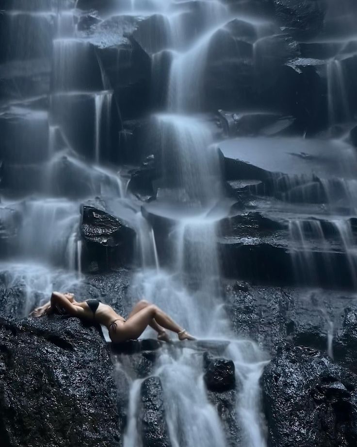 a woman laying on top of a rock next to a waterfall