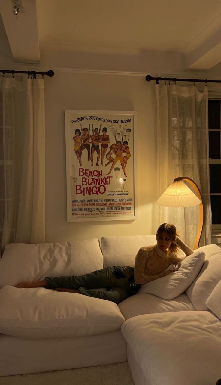 a woman laying on top of a white couch in a living room next to a lamp