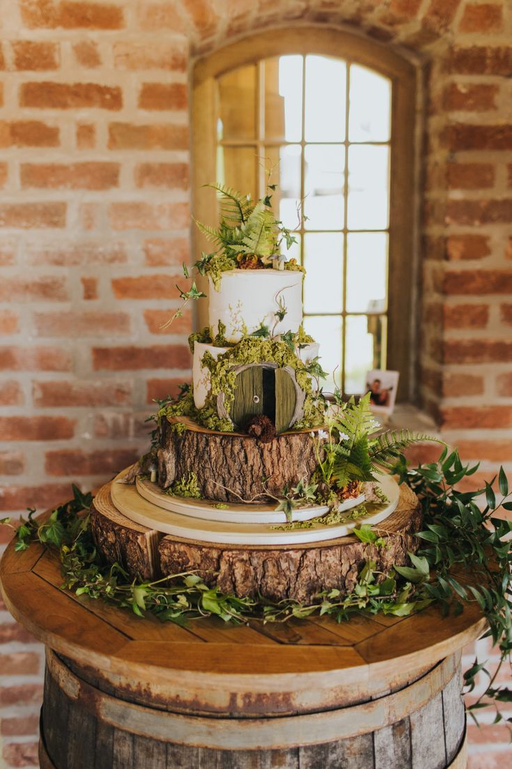 a three tiered wedding cake with greenery on top sits in front of a brick wall