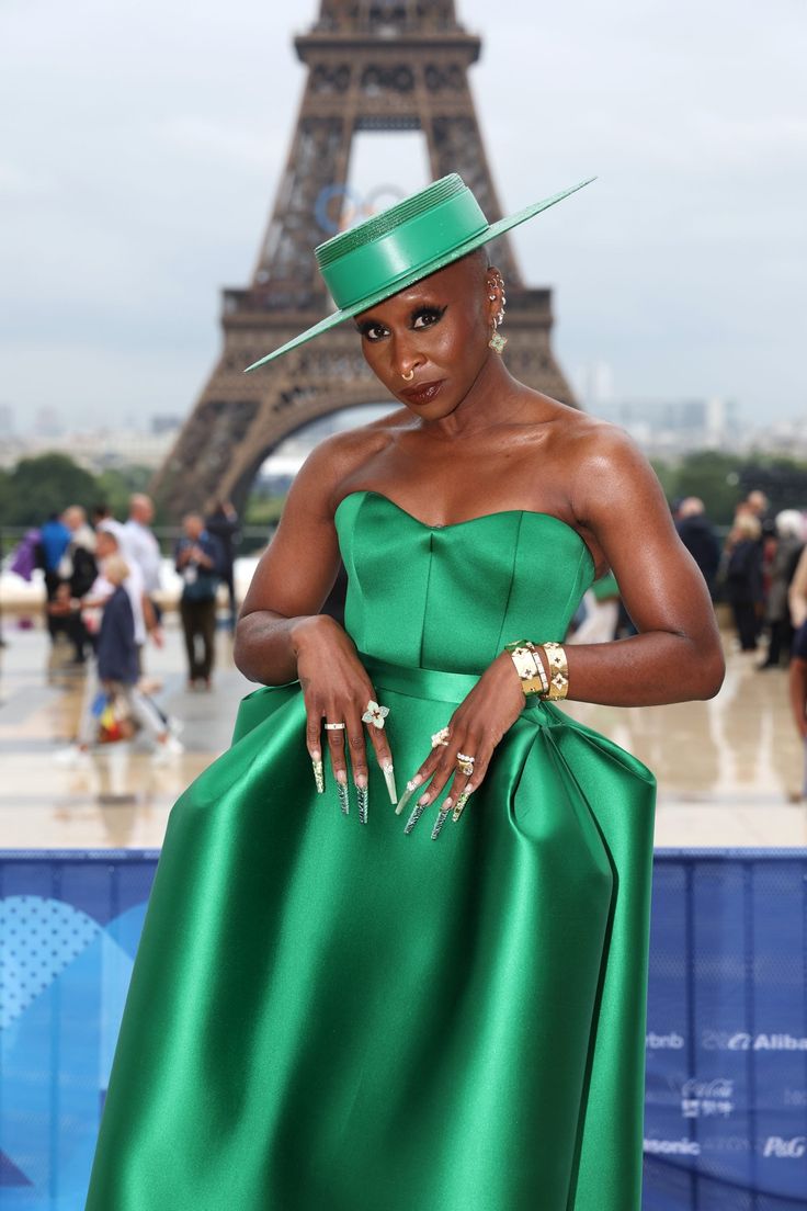 a woman in a green dress and hat poses for the camera near the eiffel tower