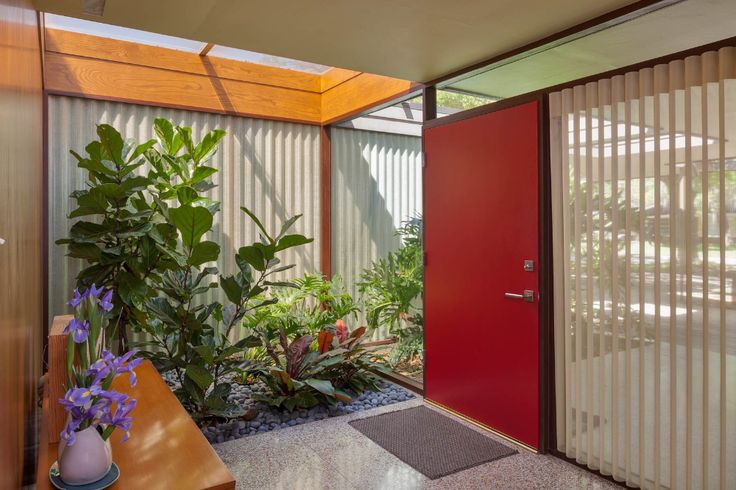 a red door is in the middle of a room with plants and flowers on the floor