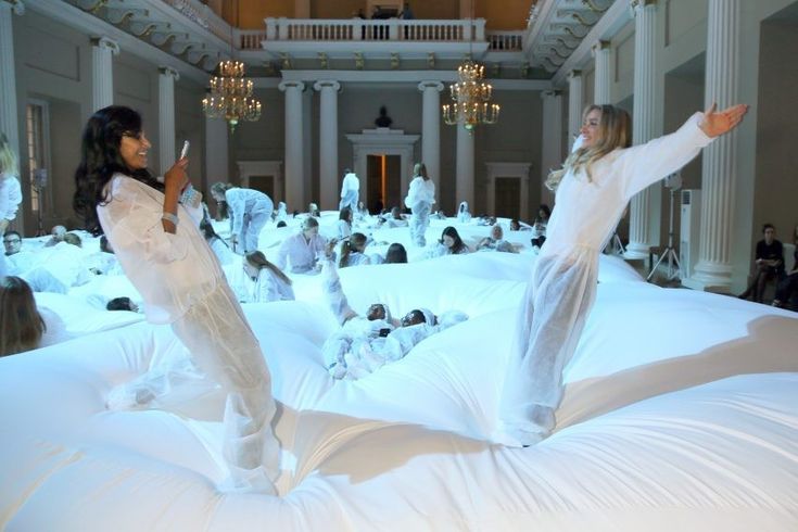 two women standing on top of a white bed in a room filled with lots of people