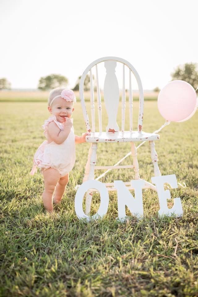 a baby girl standing next to a white chair with balloons on it and the word one written in large letters