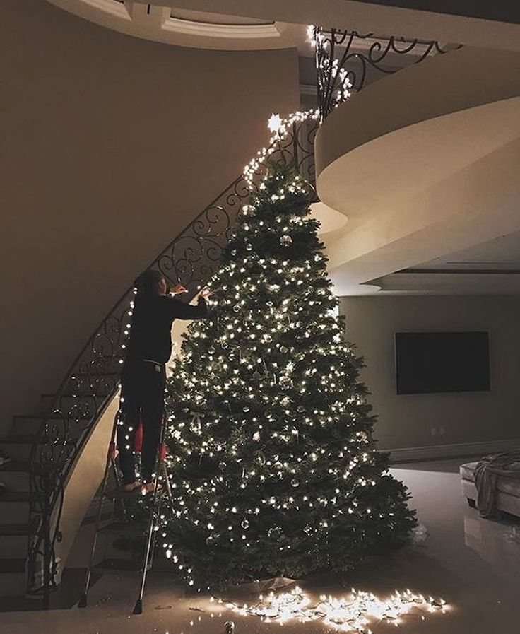 a man on a ladder next to a christmas tree in the middle of a room