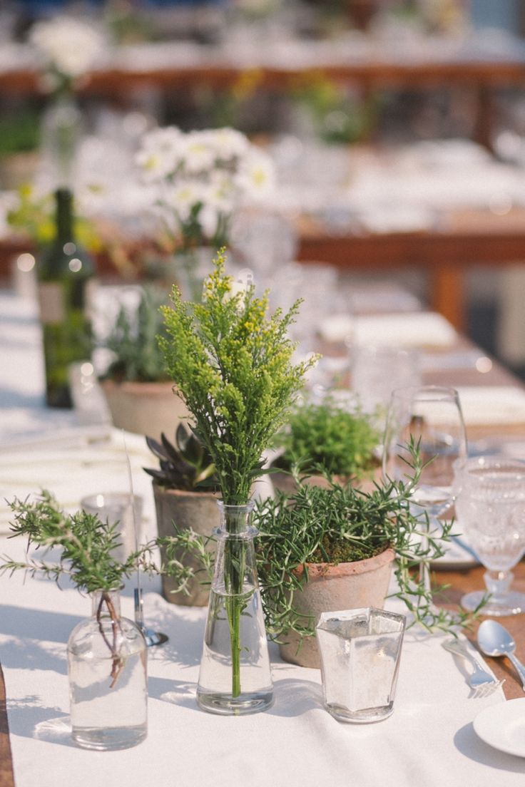 the table is set with glasses, plates and vases filled with plants on it