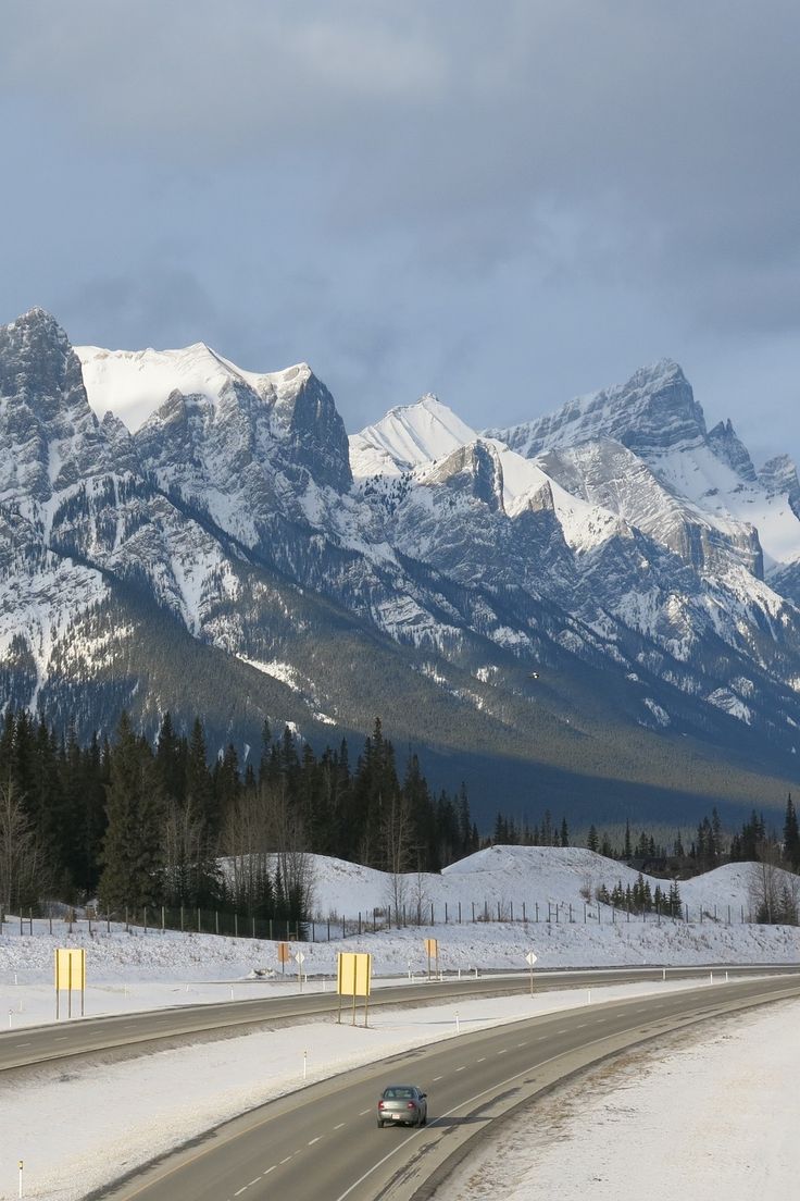 a car is driving down the road in front of mountains with snow on them and trees