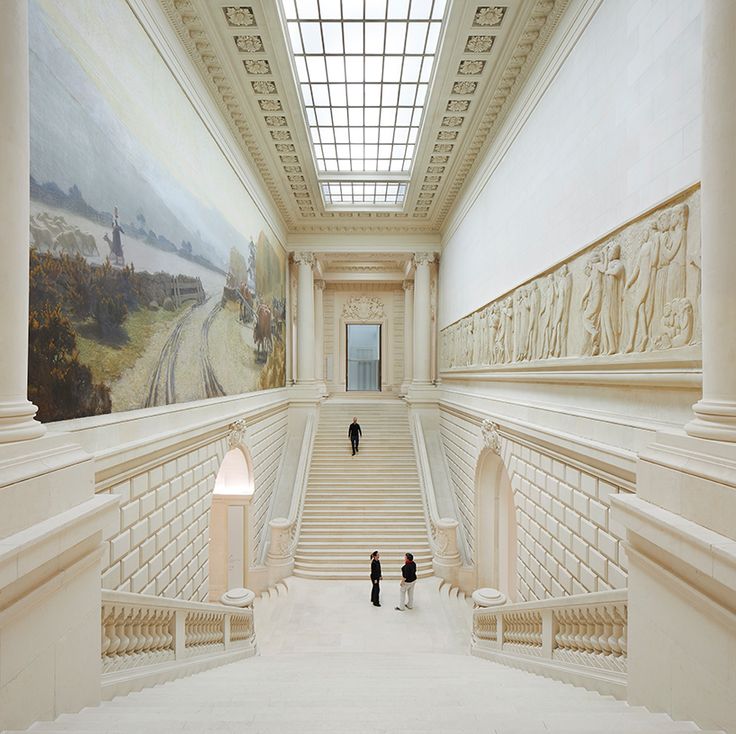 two people are standing in the middle of a large room with stairs and paintings on the walls