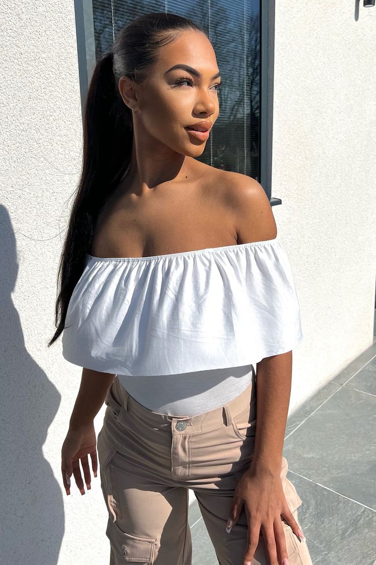 a woman with long hair standing in front of a building wearing beige pants and a white off the shoulder top