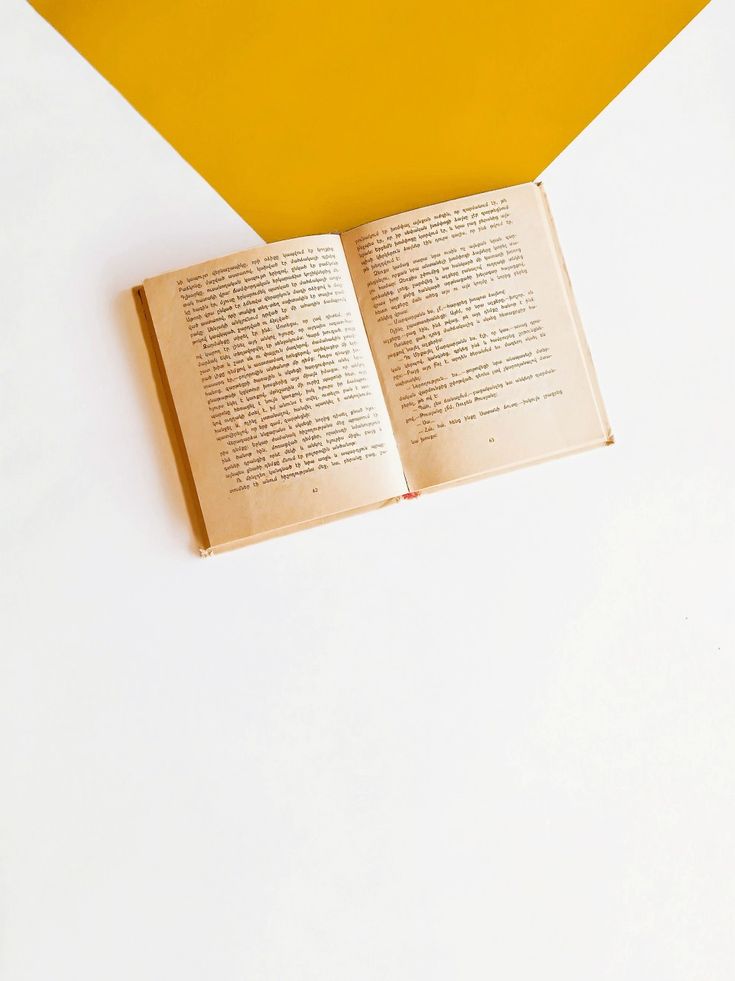 an open book sitting on top of a white table next to a yellow vase filled with flowers