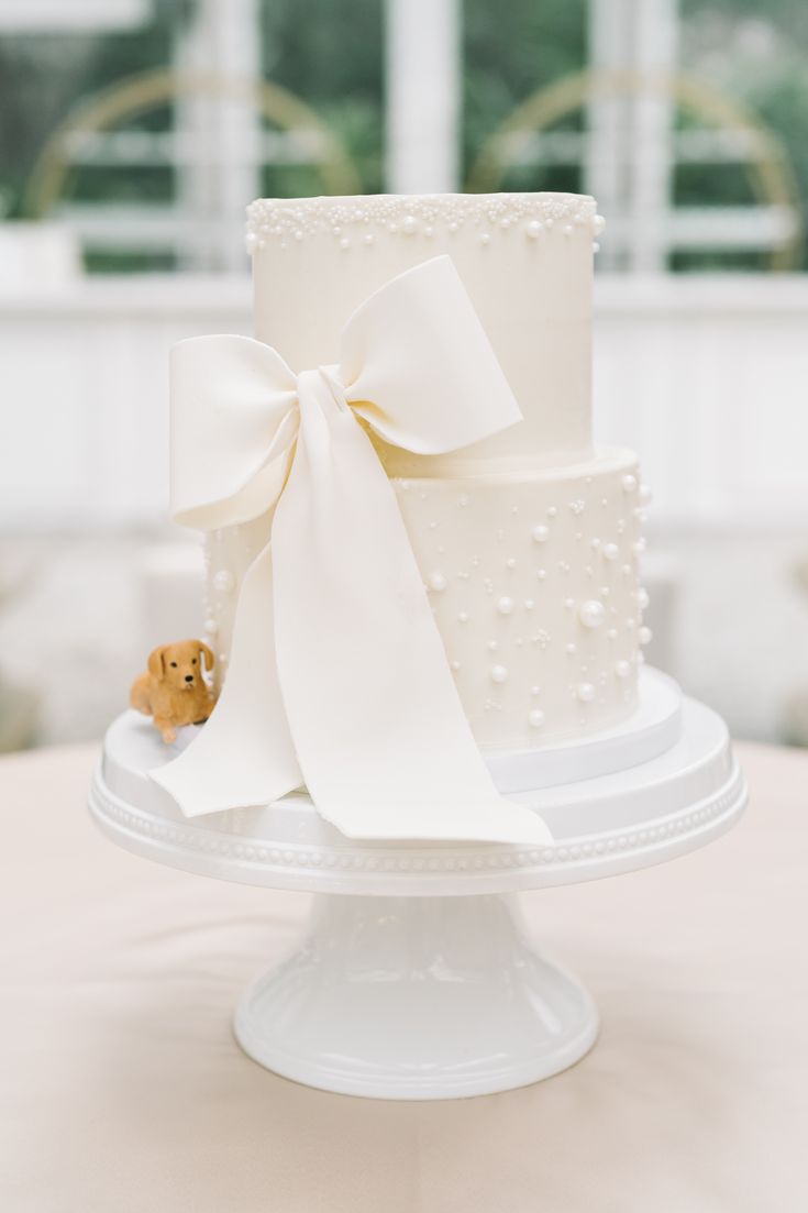 a white wedding cake with a teddy bear on the top and ribbon tied around it