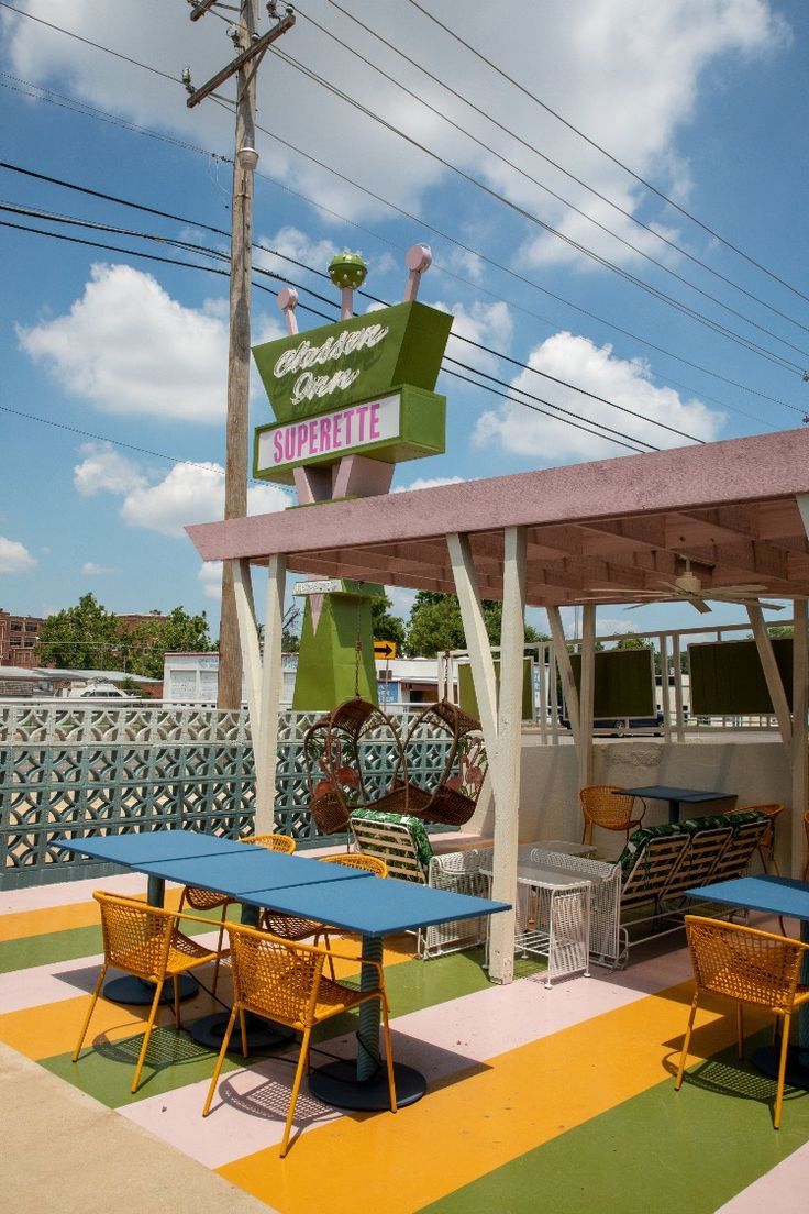 A five-photo carousel. First: a bed with a yellow spread and the wall behind it has a palm tree design with a pair of large, teal colored lips. Second: an orange, pink and green striped mural shaped like a rainbow in a guestroom. Third: the vibrant Suprette with many colors, a blue couch, pink chairs and small pieces of decor. Fourth: looking out the door from a guest room toward teal bikes under a staircase. Fifth: The patio with yellow chairs, blue tables and the hotel sign in the background. Mid Century Motel, Motel Room Design, Boutique Motel, Googie Design, Oasis Springs, Googie Architecture, Retro Motel, Motel Room, Sims Houses