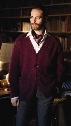 a man standing in front of a bookshelf wearing a red sweater and tie