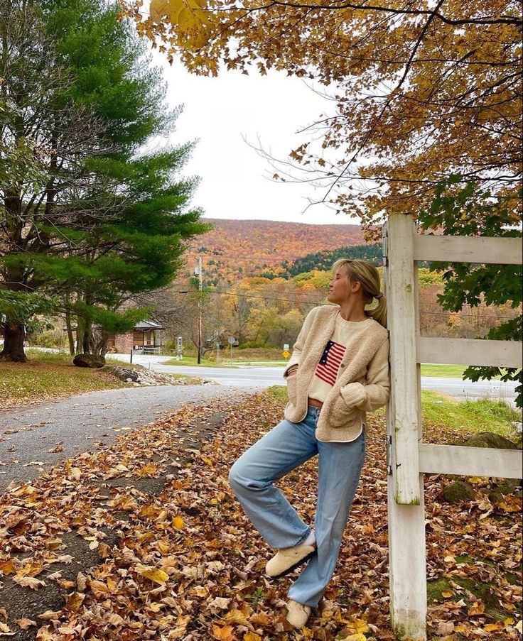 a woman leaning against a wooden fence in the fall with leaves on the ground and trees around her