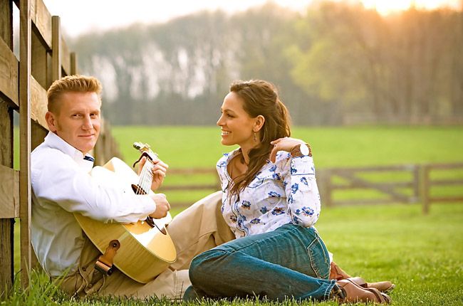 a man and woman sitting on the grass next to a fence holding an acoustic guitar