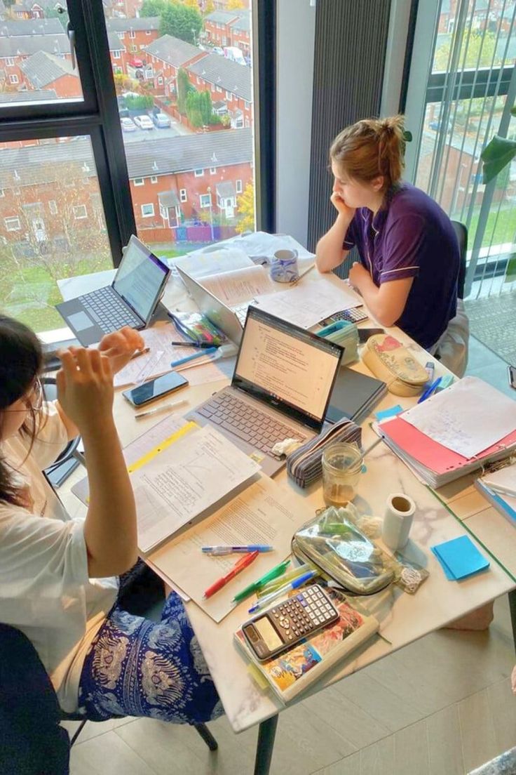 three women sitting at a table with laptops and papers on it, looking out the window