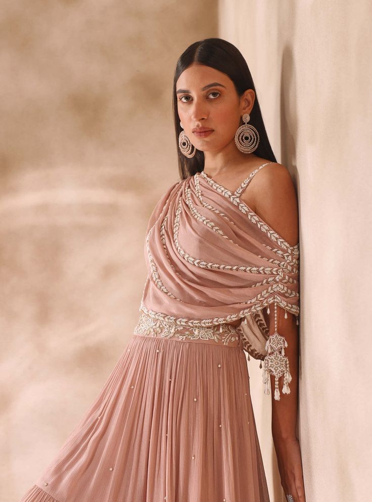 Make a style statement with our draped embellished choli featuring an attached dupatta adorned with tassels, paired beautifully with a tiered skirt delicately sprinkled with pearls. This ensemble exudes grace and charm, perfect for special occasions. Embrace the allure of traditional craftsmanship with a modern twist. Georgette Dress With Cape Sleeves For Navratri, Traditional Draped Sleeve Georgette Gown, Traditional Pink Draped Lehenga, Festive Floor-length Lehenga With Draped Sleeves, Traditional Draped Pink Lehenga, Festive Floor-length Dupatta With Draped Sleeves, Bollywood Style Dresses With Draped Sleeves, Floor-length Lehenga With Draped Sleeves For Reception, Designer Draped Gown With Sheer Dupatta