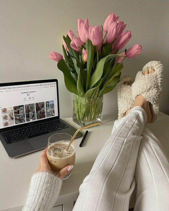 a woman holding a glass of coffee next to a laptop computer and pink tulips