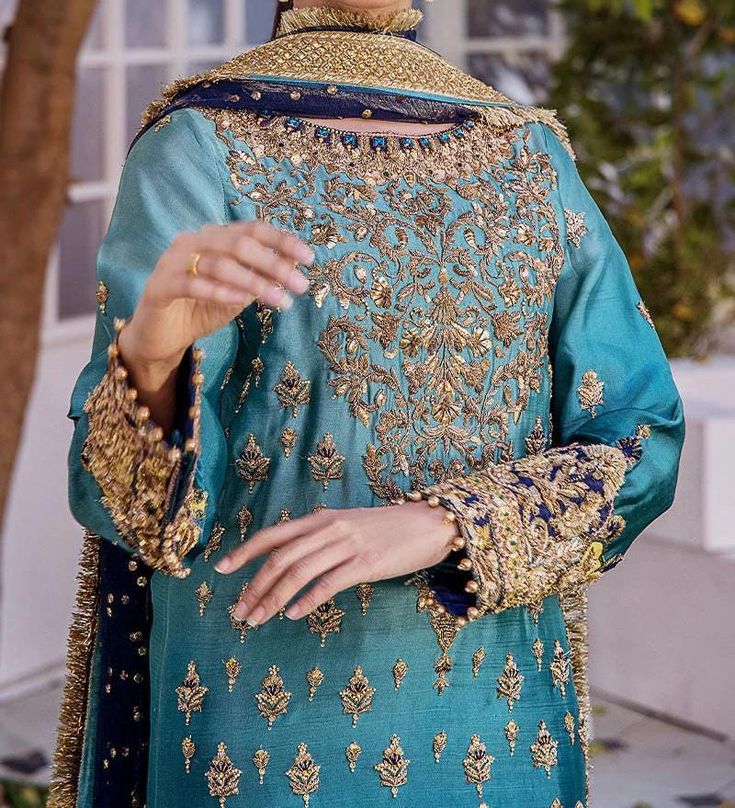 This exquisite blue wedding dress with embroidered and gold-copper work is a traditional work of art. This exquisite Kameez Trouser is the pinnacle of elegance and grace thanks to its flawless stitching and embroidered borders. The stunning kameez features hand-crafted zardosi and gold-copper accents, and it has an enticing shade of blue. This kameez is ideal for a wedding because of the embroidery on the front and around the borders. This elaborately embroidered kameez is worn as a magnificent wedding dress when coupled with tissue-fabric trousers. The pants' edges are embellished with goldwork and Gold Embroidered Raw Silk Dress, Blue Embroidered Salwar Kameez For Wedding, Gold Embroidered Sharara For Reception, Gold Embroidered Fabric For Eid Reception, Gold Jamawar Sharara For Reception, Gold Embroidered Jamawar Dress, Eid Gold Embroidered Raw Silk Fabric, Blue Jamawar Wedding Dress, Gold Brocade Dress With Resham Embroidery