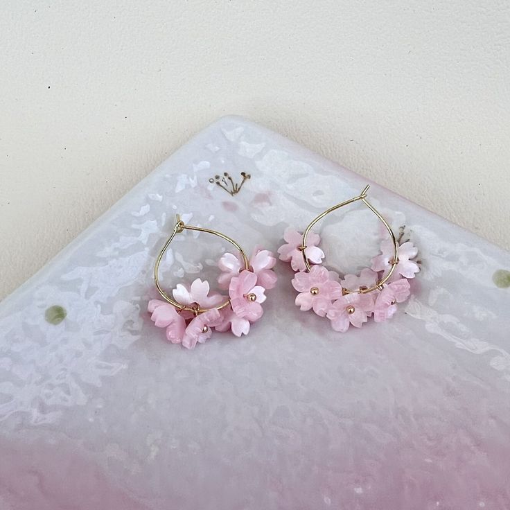 These exquisite hoop earrings feature delicate cherry blossom sakura flowers, available in either pink or white hues.  Each earring is meticulously handcrafted and the ear hoops are plated with  14K gold, exuding elegance and charm.  Perfect for adding a touch of whimsy and sophistication to any outfit, these earrings are versatile enough to be worn as bridal accessories or to elevate everyday looks with a dash of kawaii (cuteness). Hoop Flower Charm Earrings As Gift, Dainty Spring Flower Earrings For Pierced Ears, Spring Hoop Earrings With Flower Charm As Gift, Spring Flower Charm Hoop Earrings As Gift, Spring Gift Hoop Earrings With Flower Charm, Dainty Hoop Flower Earrings Gift, Handmade Delicate Flower Hoop Earrings, Spring Flower Hoop Earrings For Pierced Ears, Small Hoop Pink Flower Earrings