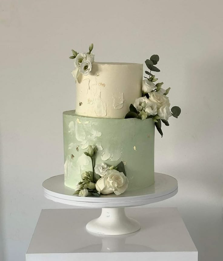 a three tiered cake with white flowers and greenery on the top is sitting on a pedestal