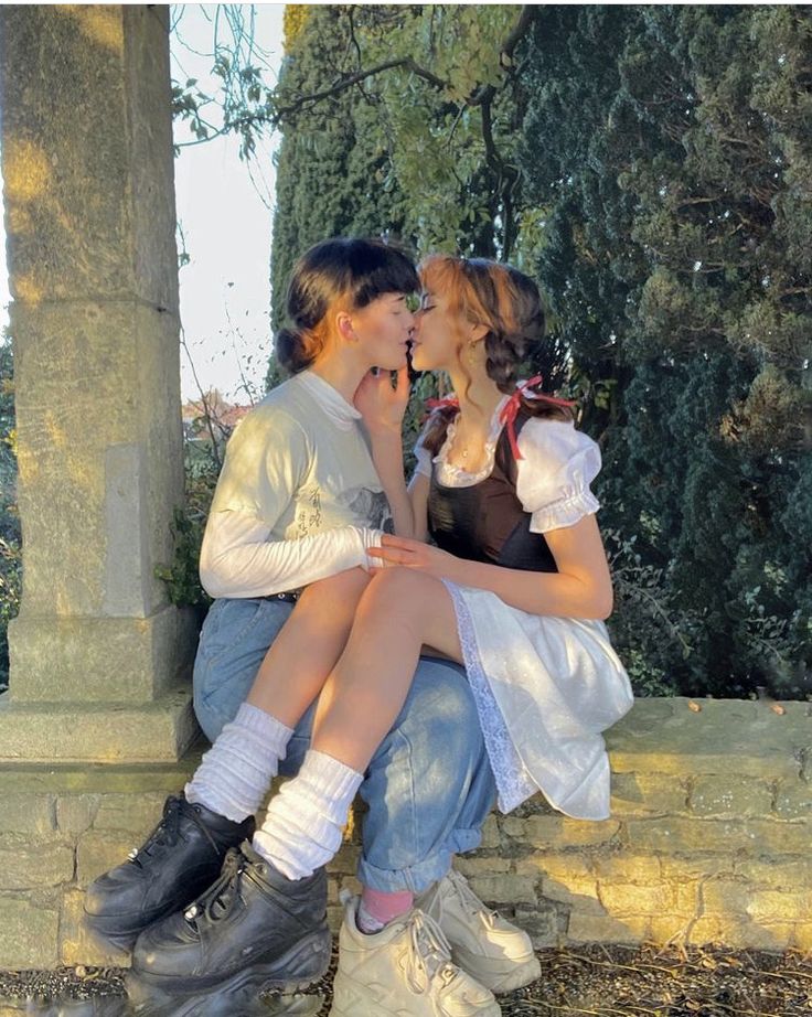 two people sitting on top of each other in front of a stone wall and trees