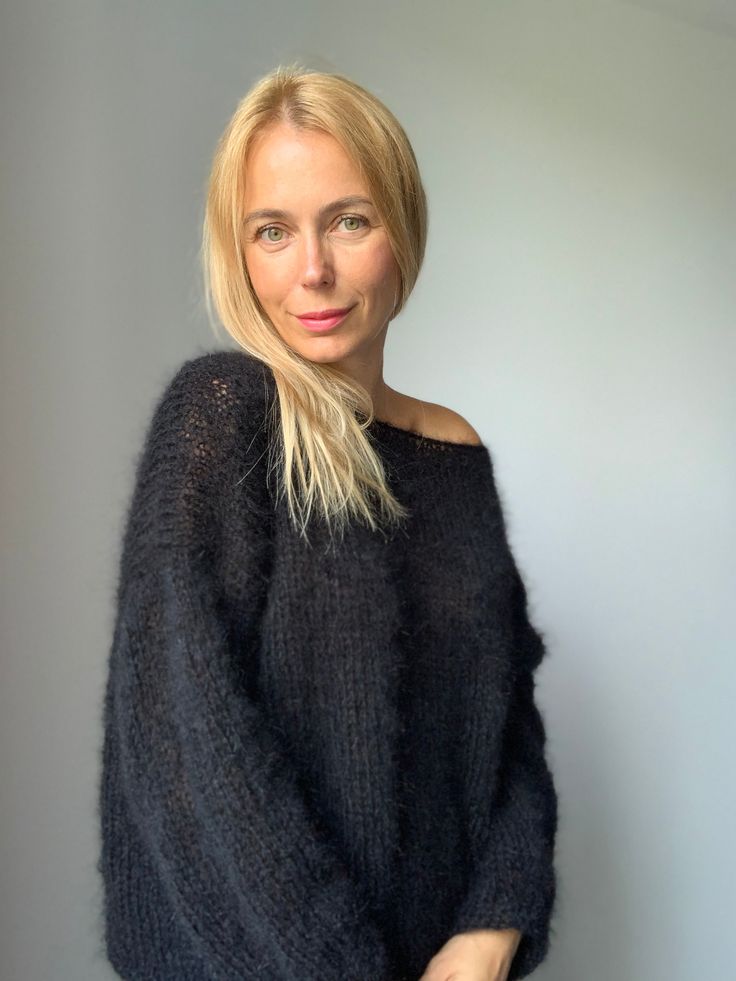 a woman standing in front of a white wall wearing a black sweater and blue jeans