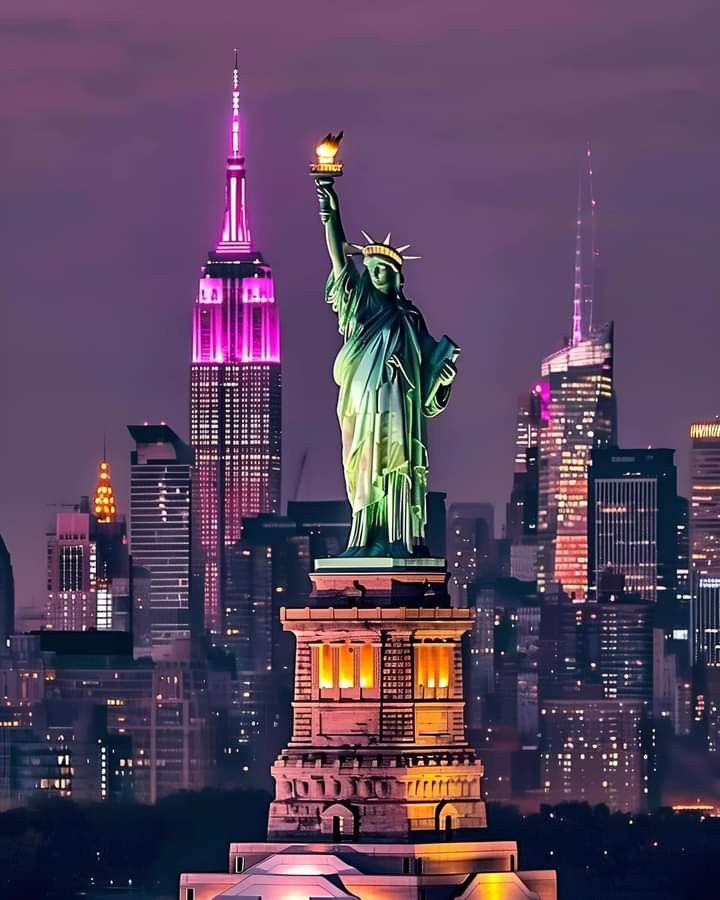 the statue of liberty is lit up in pink and purple colors with city lights behind it