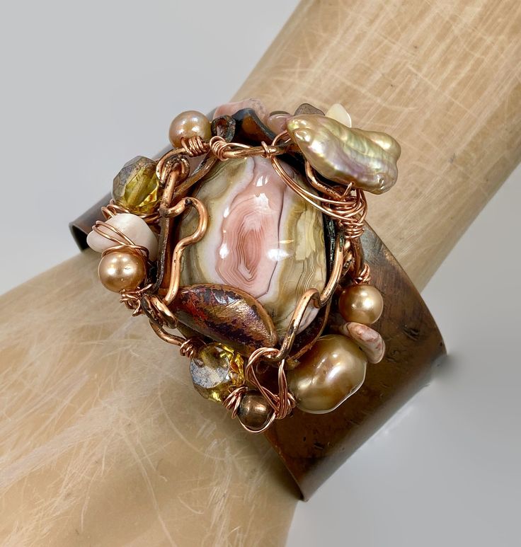 "Pink and shades of soft browns and beige....  Starting with a copper cuff bracelet that I have hammered and heated for an amazing color... I then chose the large jasper stone that is the centerpiece of this bracelet. I love the swirling shades of pink and cream.  I encased it in a freeform, forged copper bezel with a unique organic shape to it.  I have given it an artistic and organic wire wrap . For additional accents I have used beads in stone, glass and pearls to enhance the shades of the st Brown Hand Wrapped Cuff Bracelet For Gift, Unique Bronze Wire-wrapped Bracelets, Unique Wire Wrapped Bronze Bracelets, Unique Bronze Wire Wrapped Bracelets, Adjustable Brown Copper Bangle, Bohemian Copper Bangle For Gifts, Bohemian Copper Bangle As A Gift, Bohemian Adjustable Copper Bracelets, Bohemian Hand Wrapped Brown Bracelets