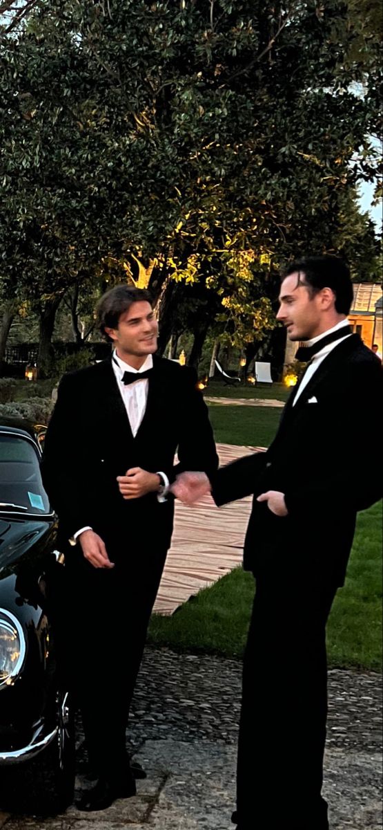 two men in tuxedos are shaking hands next to a black car on the street