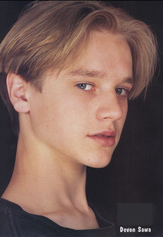 a young man with blonde hair and blue eyes looks into the camera while wearing a black t - shirt