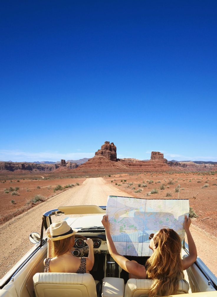 two women sitting in the back of a convertible car looking at a map while on a desert road