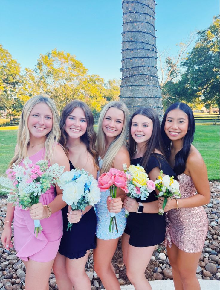 four beautiful young women standing next to each other in front of a palm tree holding bouquets