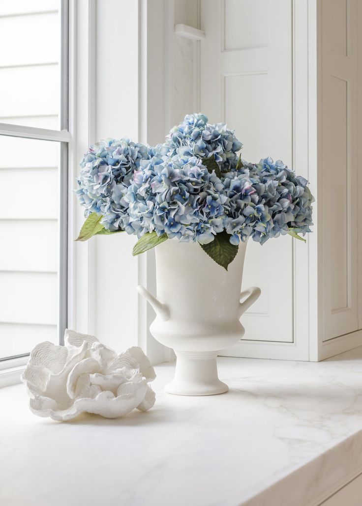 a white vase filled with blue flowers sitting on top of a counter next to a window