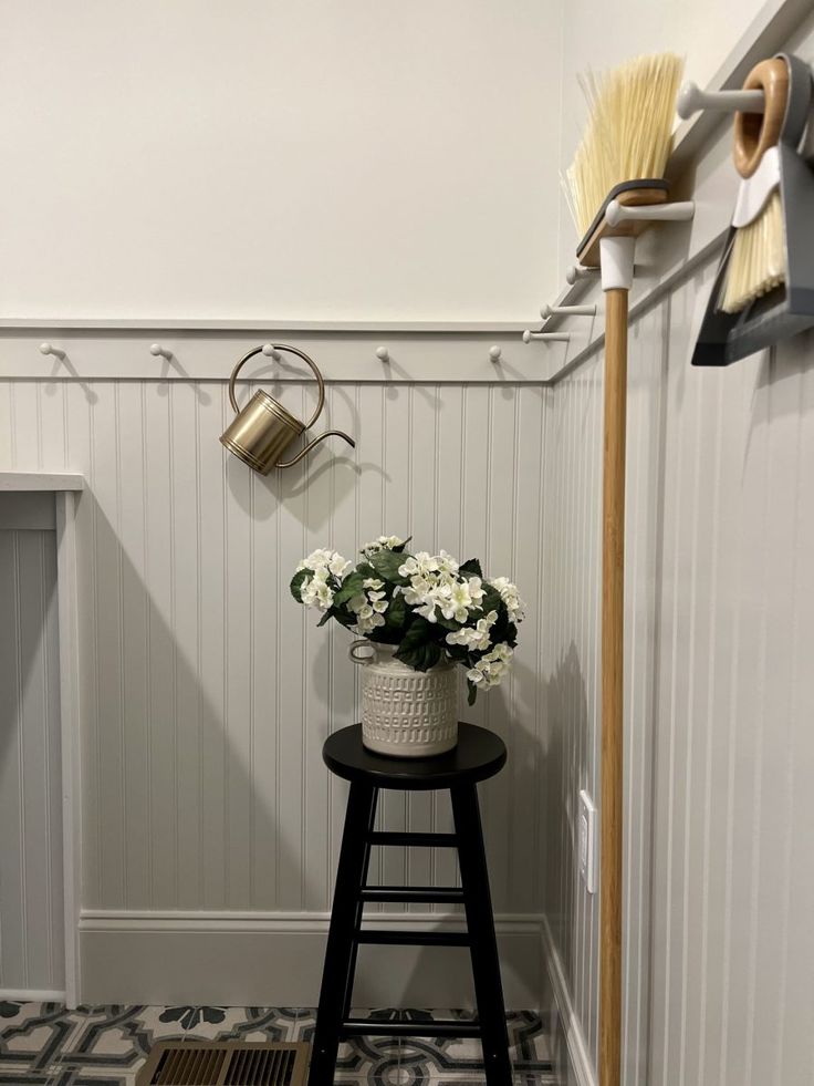 a vase with flowers sitting on top of a stool next to a wall mounted broom