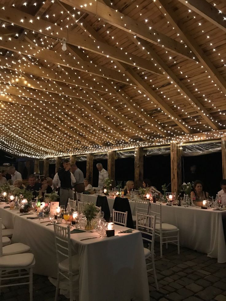 people are sitting at tables with white linens and string lights on the ceiling above them