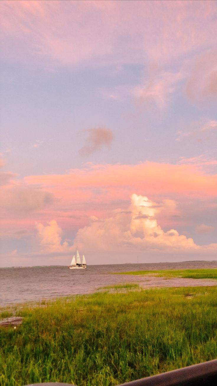 the sailboat is sailing in the distance on the water near the grass and trees