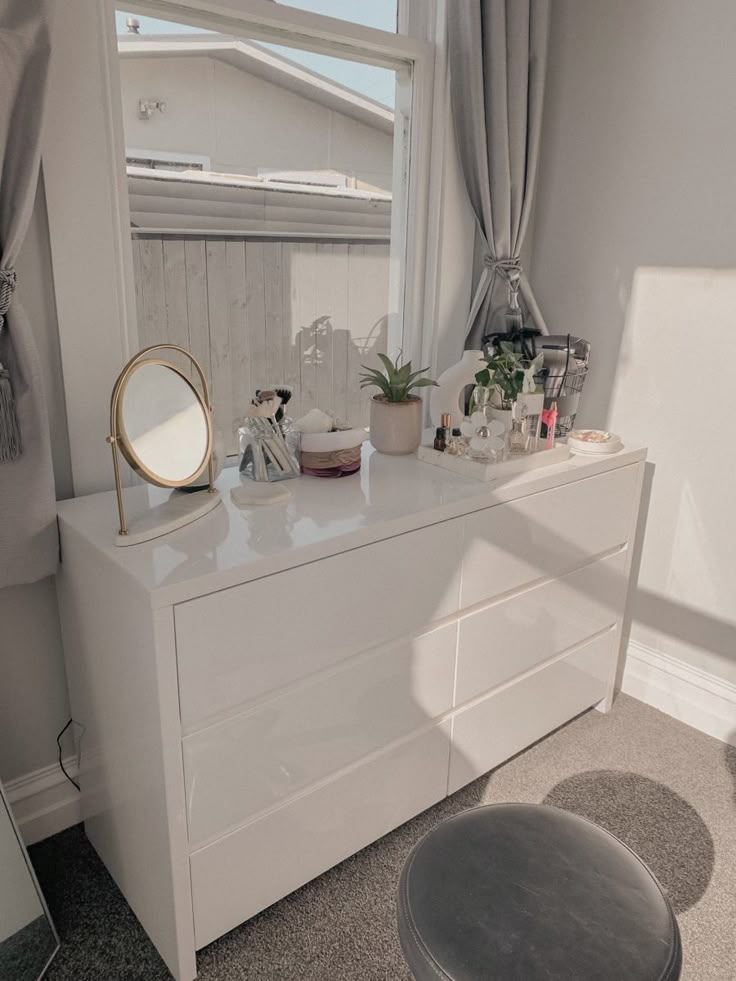 a white dresser sitting in front of a window next to a chair and table with a mirror on it