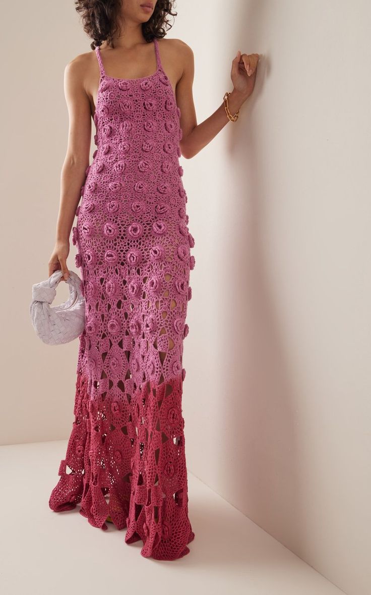 a woman in a pink crochet dress leaning against a wall holding a white purse