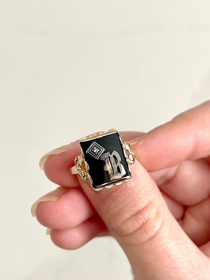 STUNNING bold and regal vintage yellow gold ring with a rectangular black onyx stone in the center and gorgeous scalloped ornate detailing beneath the gold pedestal holding the onyx up. The onyx bears a silver letter "B" on top with a small but mighty diamond stone in the corner. This is an incredible, highly sought-after art deco signet ring. This is estimated to be an antique piece. There is a symbol stamped inside the band beside which I cannot make out. This is presumably the maker's mark but possibly also the gold content. It appears to be a triangle, which apparently has roots in England and may even indicate a higher gold content, such as 14K. Size is approximately 6 - 6.25. Total weight is approximately 2.59 grams. *Note: This listing will ship with TRACKING to ALL destinations, in Signet Ring With Diamond, Gold Pedestal, B Initial, Onyx Signet Ring, Black Onyx Stone, Ring With Diamond, Letter B, Scalloped Edges, Maker's Mark