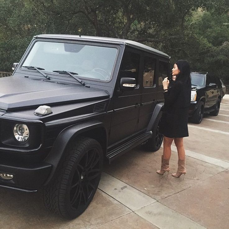 a woman standing next to a black mercedes g - class parked in a parking lot