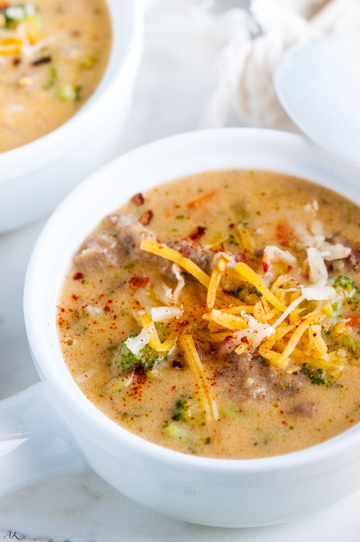 two bowls of soup with cheese and broccoli in them on a white table