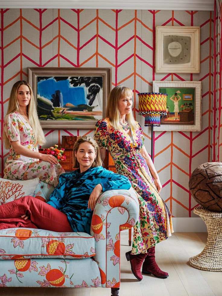 two women sitting on a couch in front of a wallpapered room with paintings