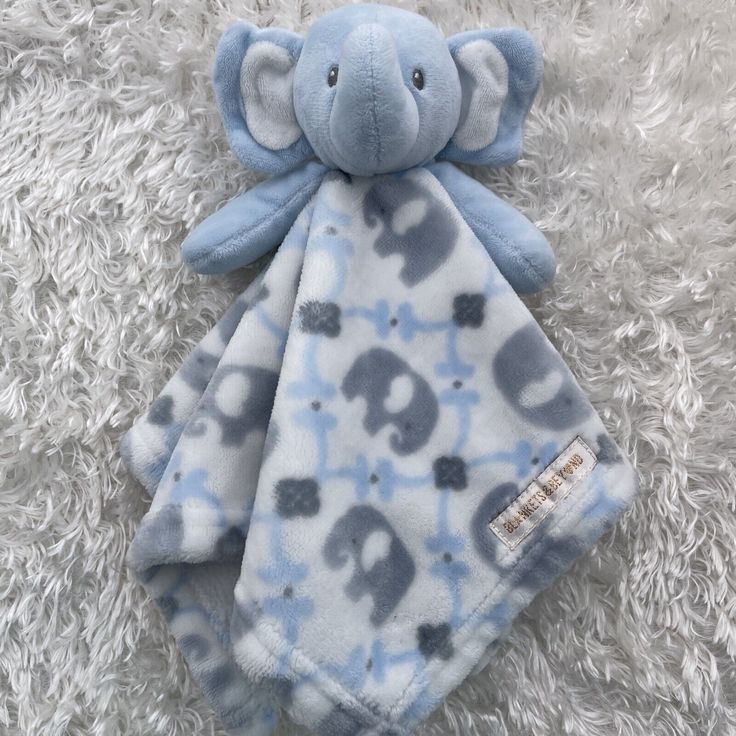 a blue and gray elephant blanket laying on top of a fluffy white carpet next to a stuffed animal