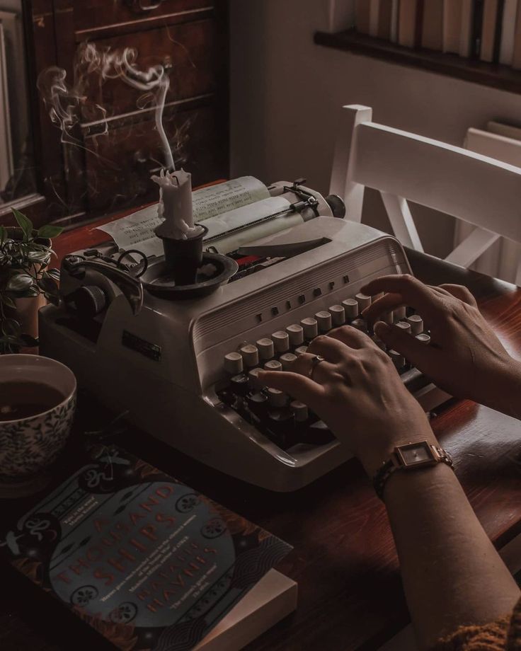 a person typing on an old fashioned typewriter at a table with books and cups